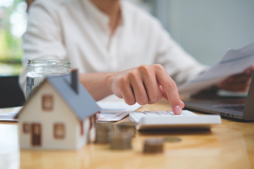Pessoa utilizando calculadora para simular financiamento imobiliário com modelo de casa e moedas sobre a mesa.