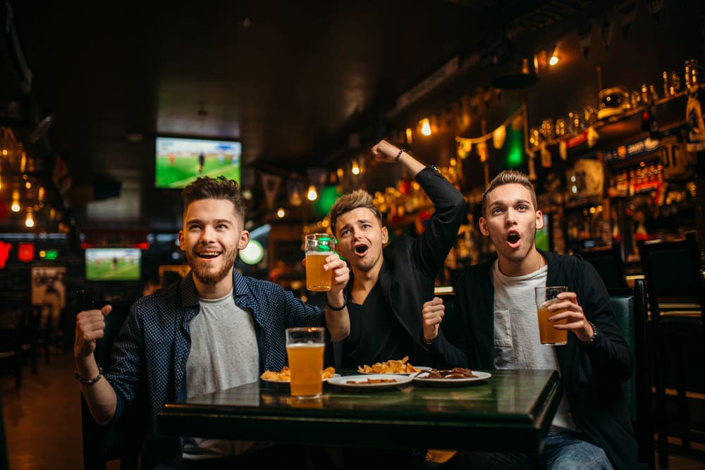 bar com telão e três amigos assistindo a uma partida de futebol, enquanto bebem cervejas