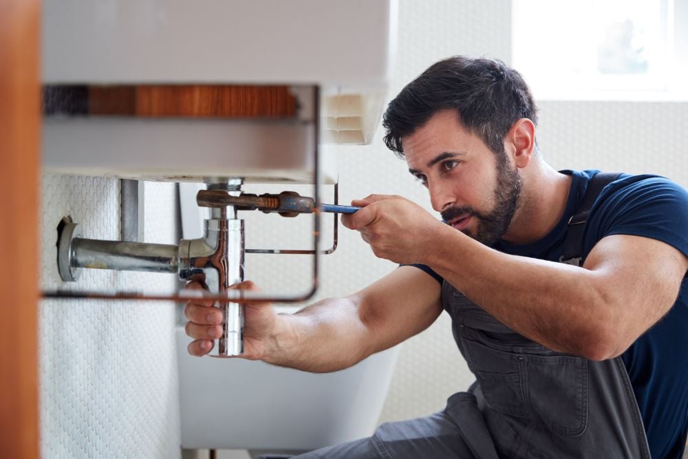 Profissional de manutenção utiliza uma chave de grifo para ajustar a tubulação de metal sob uma pia, demonstrando uma etapa de como tirar ar do encanamento.