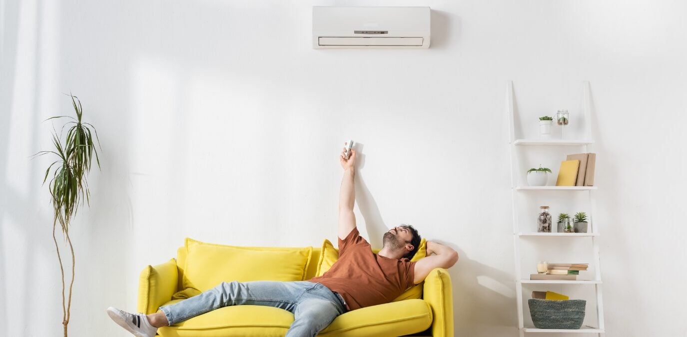 Homem relaxa no sofá amarelo controlando ar-condicionado enquanto calcula btus metros quadrados para conforto ideal.