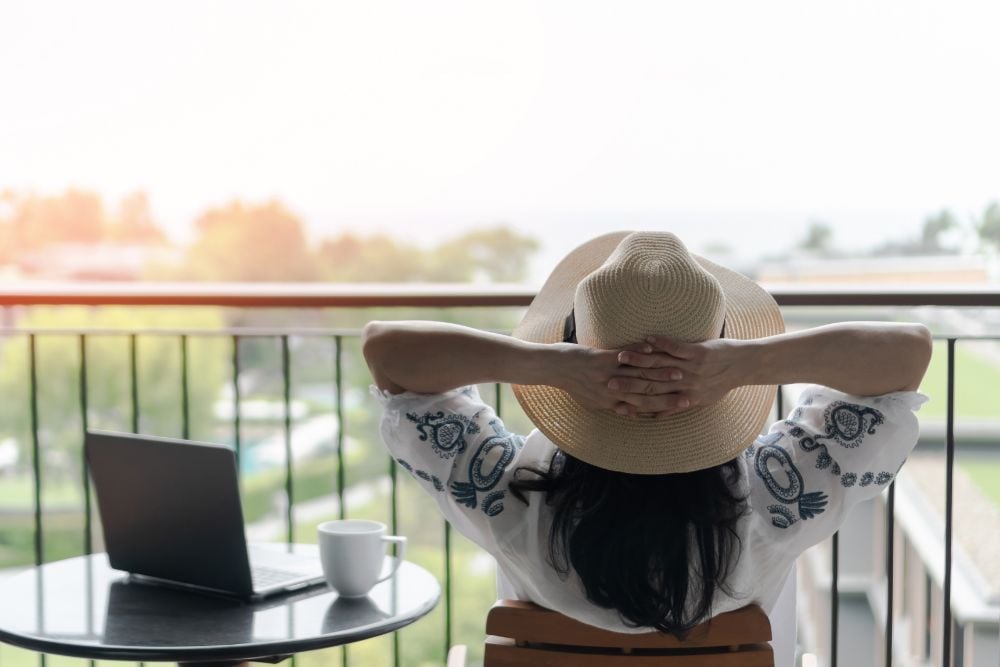 Mulher relaxando na varanda com notebook e café, aproveitando um momento de tranquilidade e bem-estar que reflete uma boa qualidade de vida.