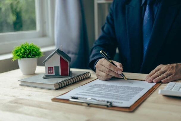 Corretor preenchendo contrato com miniatura de casa sobre a mesa, simbolizando o seguro fiança na prática para aluguel de imóveis.