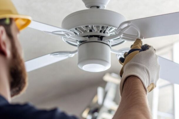 Eletricista com capacete e luvas brancas montando uma das pás brancas do ventilador, mostrando o passo a passo de como instalar ventilador de teto