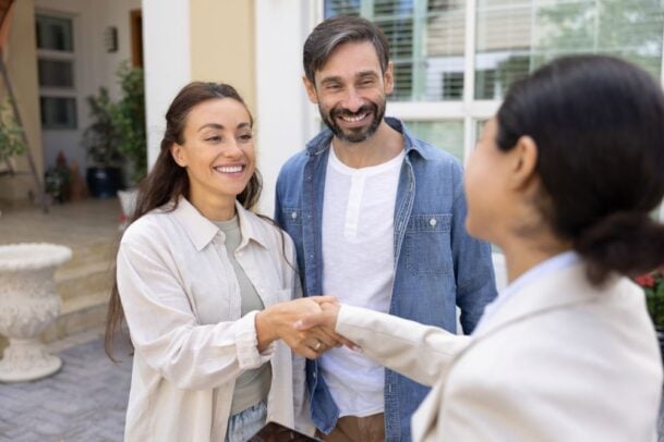Caução aluguel vantagens e desvantagens: Casal feliz apertando a mão de uma corretora em frente a uma casa, finalizando o contrato de aluguel.