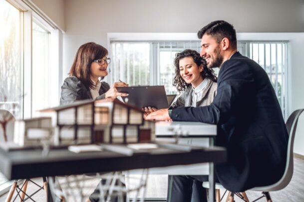 Casal em uma mesa de vendas analisando a maquete de um novo empreendimento e conversando com uma corretora, exemplificando o processo de compra de um lançamento de imóvel.