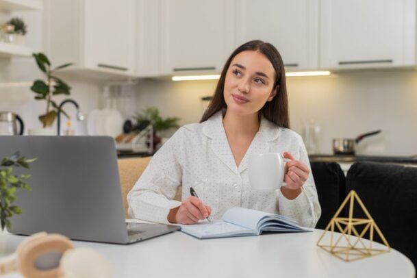 Mulher planejando a lista de chá de cozinha com um caderno e caneta na mesa, tomando café em uma cozinha clara.