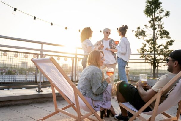 Jovens sentados em cadeiras de madeira relaxando em um terraço decorado com varal de luzes durante o pôr do sol, exemplificando na prática o que é rooftop.