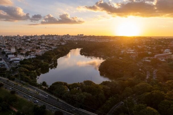 Imagem aérea do parque taquaral, com destaque para o lago e pôr do sol ao fundo, uma das principais opções do que fazer em Campinas para casais.