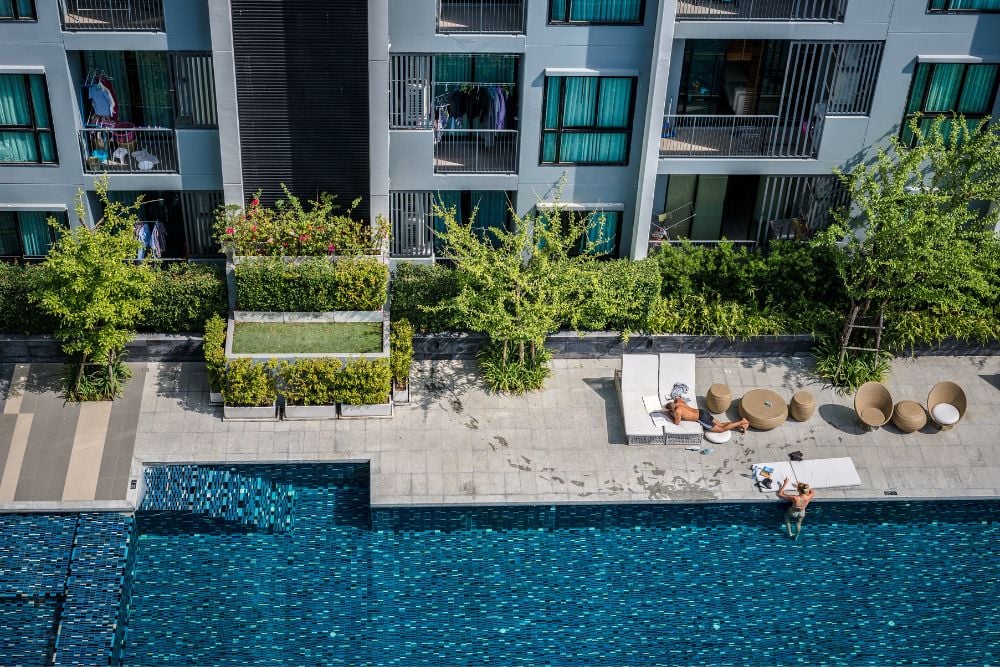 Área da piscina moderna em um condomínio clube, com pessoas relaxando em espreguiçadeiras e aproveitando o sol próximo à fachada do edifício.