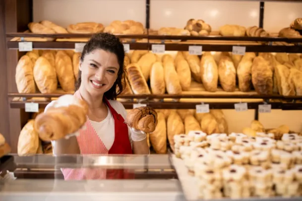 Imagem de uma mulher atendente de padaria mostrando alguns pães, representando a qualidade de atendimento nas padarias da Vila Nova Conceição.