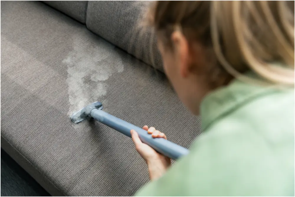 Pessoa higienizando sofá de tecido com vaporizador, ilustrando métodos eficientes de como posso limpar um sofá de tecido e remover sujeiras profundas.