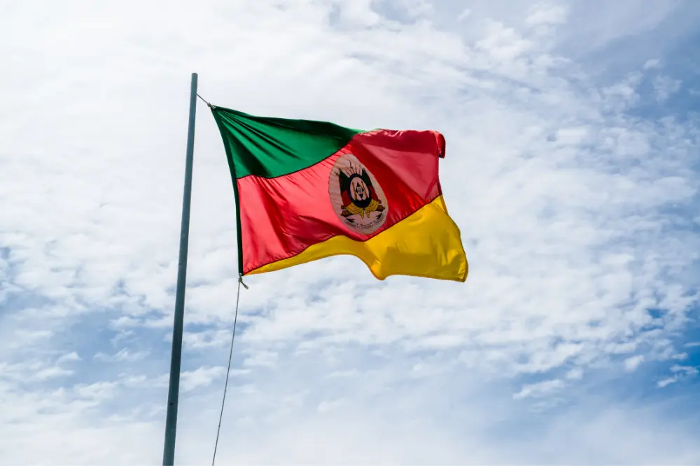 Bandeira do Rio Grande do Sul tremulando ao vento em céu azul com nuvens, representando o estado no contexto do ranking das maiores cidades do-RS.
