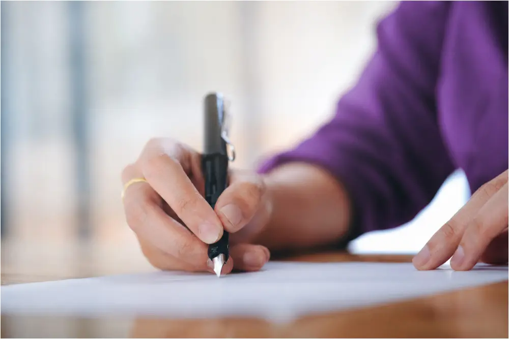 Pessoa assinando contrato imobiliário em mesa de madeira, representando processo de cessão de direitos e responsabilidades do cessionário em negociações imobiliárias.