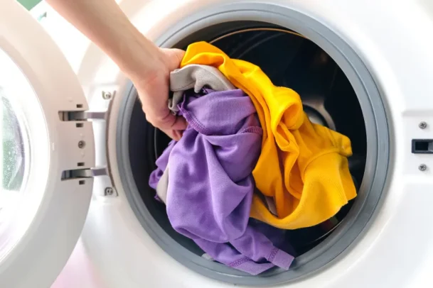 Pessoa colocando roupas coloridas na lavadora, ilustrando cuidados básicos e práticas recomendadas sobre como limpar máquina de lavar regularmente em casa.